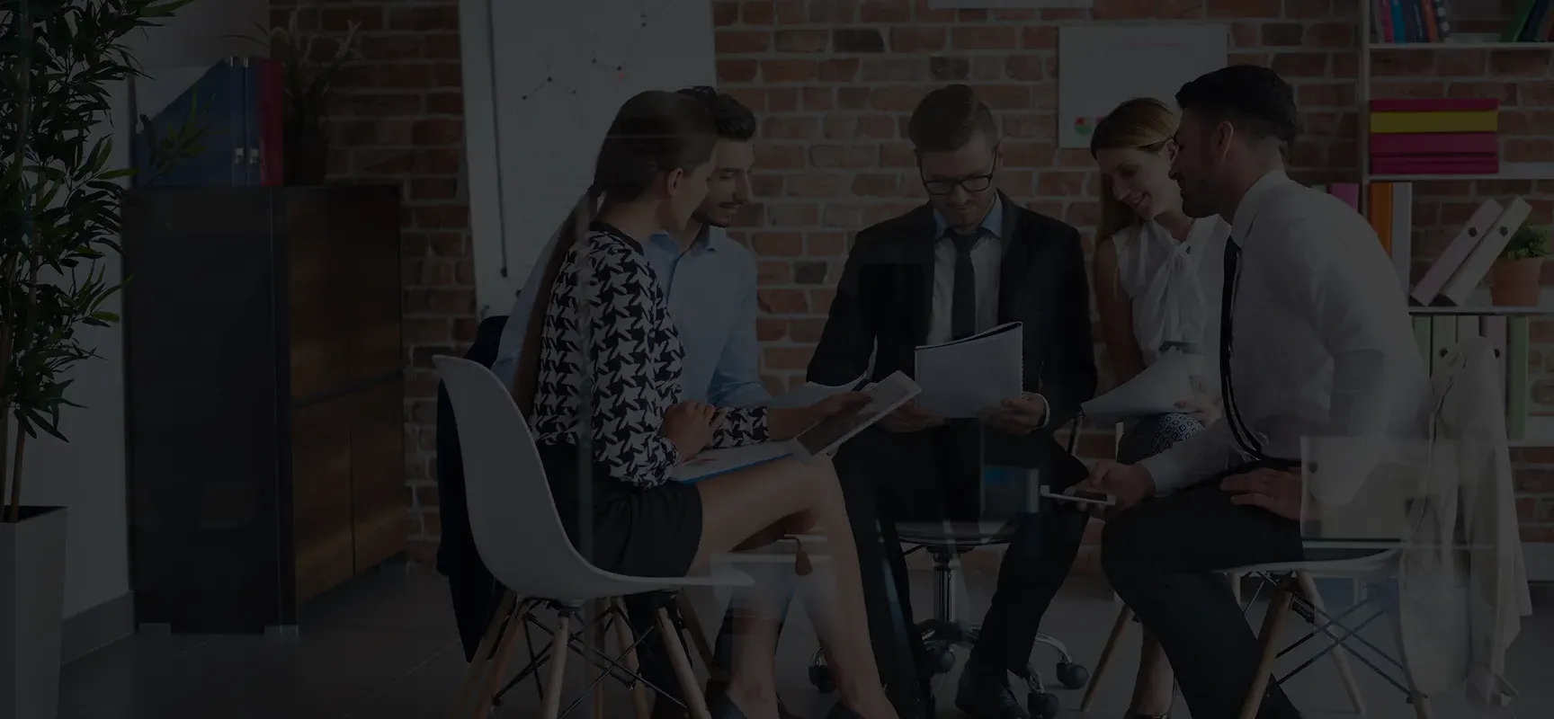 Five professionals in business attire seated in a modern office, discussing documents in a collaborative meeting.
