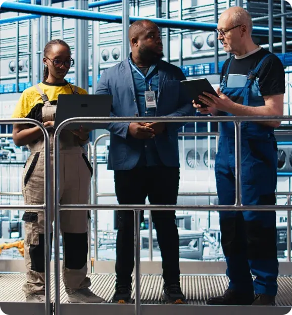 Three industrial workers in a factory discussing work, one holding a laptop, another a tablet.