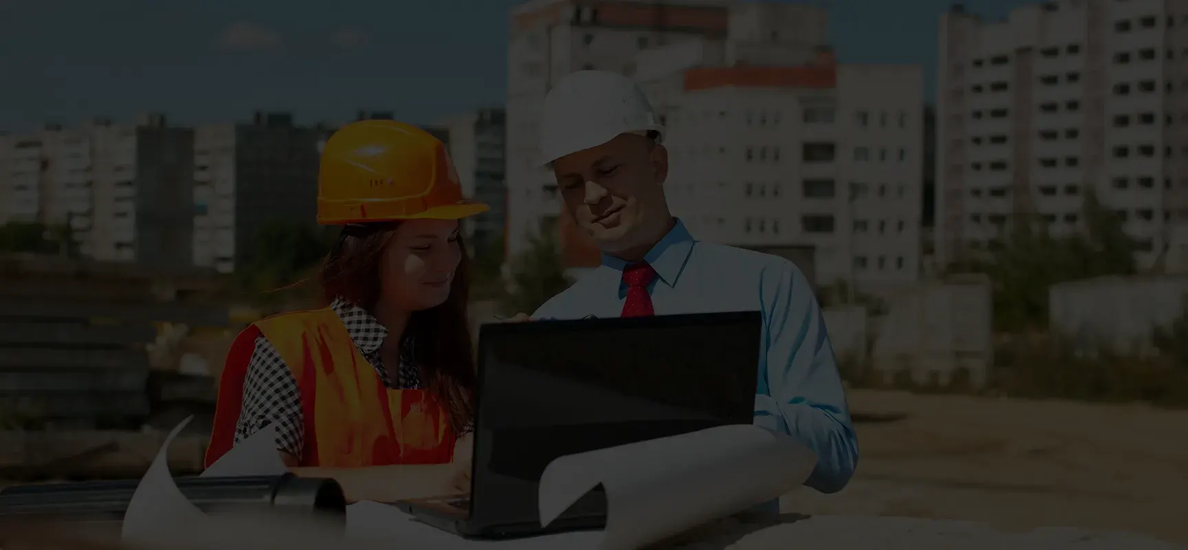 Two construction professionals reviewing plans on a laptop at a building site with urban apartment buildings in the background.