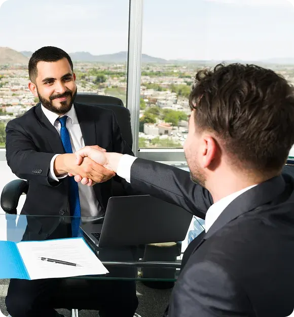 Two businessmen in suits shaking hands across a glass desk with a laptop and documents, cityscape visible through the window.