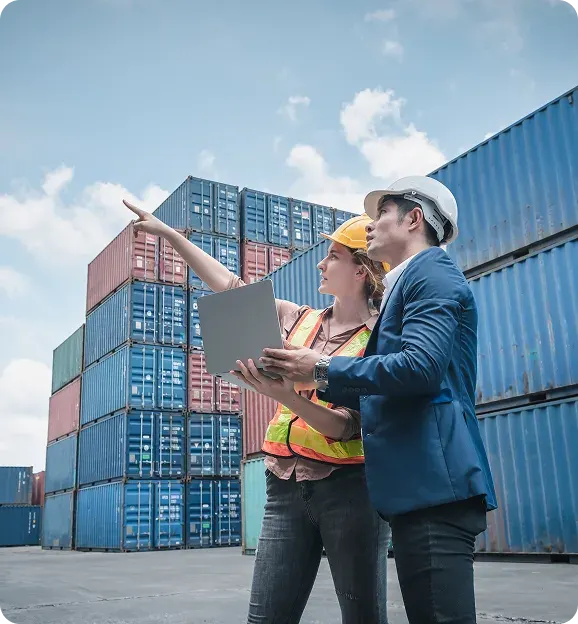Two workers in safety helmets and vests inspecting stacked cargo shipping containers, one holding a laptop and the other pointing.