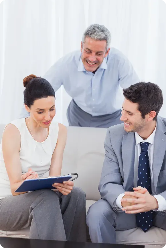 Three business professionals seated on a sofa reviewing a document on a clipboard, smiling and engaged in discussion.