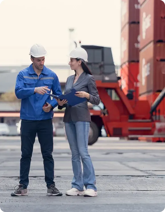 Man and woman wearing white safety helmets reviewing documents in an industrial shipping container yard.