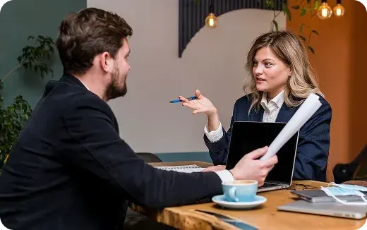 Two business professionals having a discussion across a table with a laptop, documents, and a coffee cup.