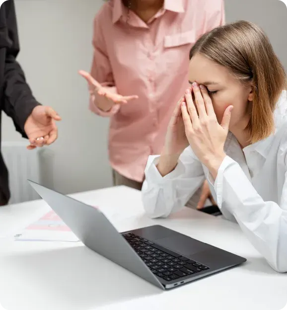 Stressed woman sitting at a desk with a laptop, covering her face with her hands, while two people in the background gesture expressing frustration.