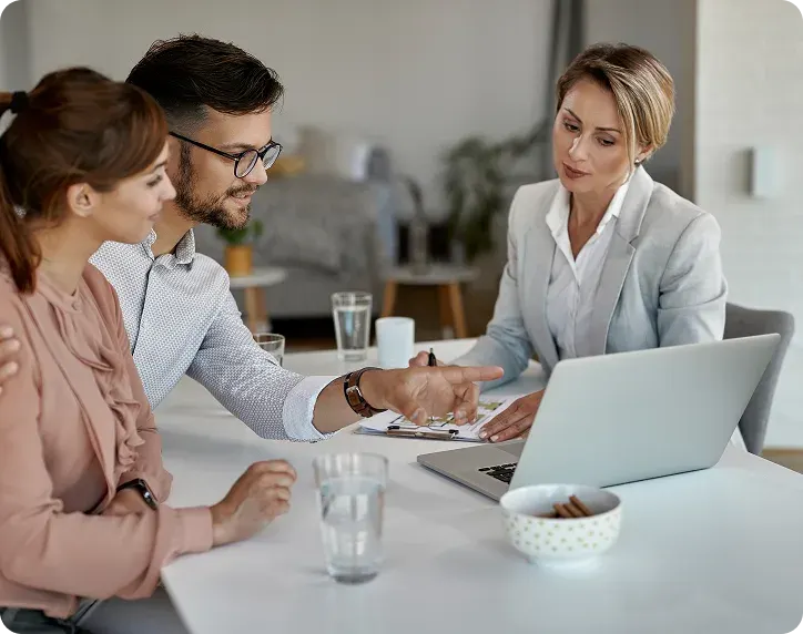 Two people sitting at a table discussing with a businesswoman who is using a laptop and holding a pen.
