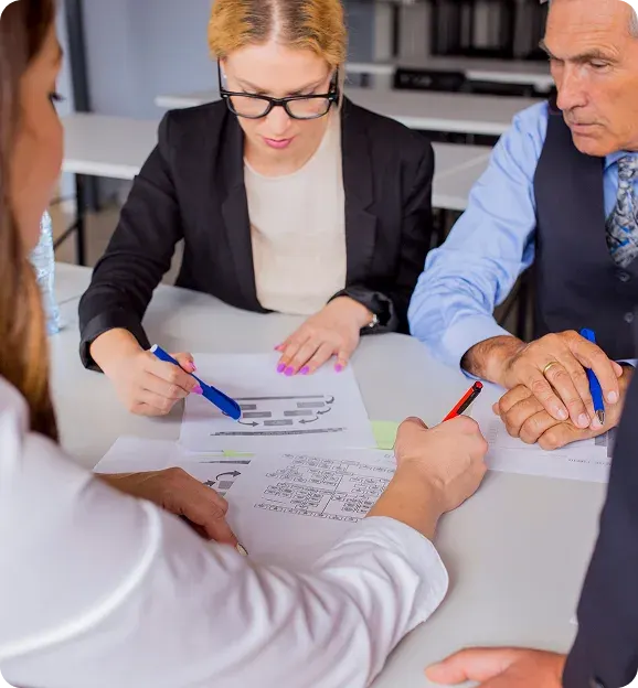 Three people seated around a table reviewing and marking documents and charts with pens during a meeting.