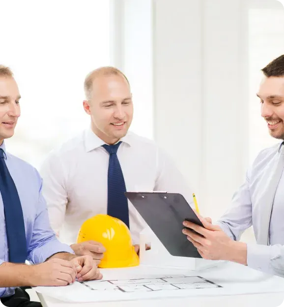 Three men in business attire reviewing architectural plans on a table, with a yellow hard hat and a clipboard.