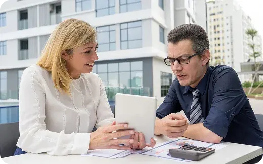 Two business professionals discussing documents and a tablet at an outdoor table with a city building background.