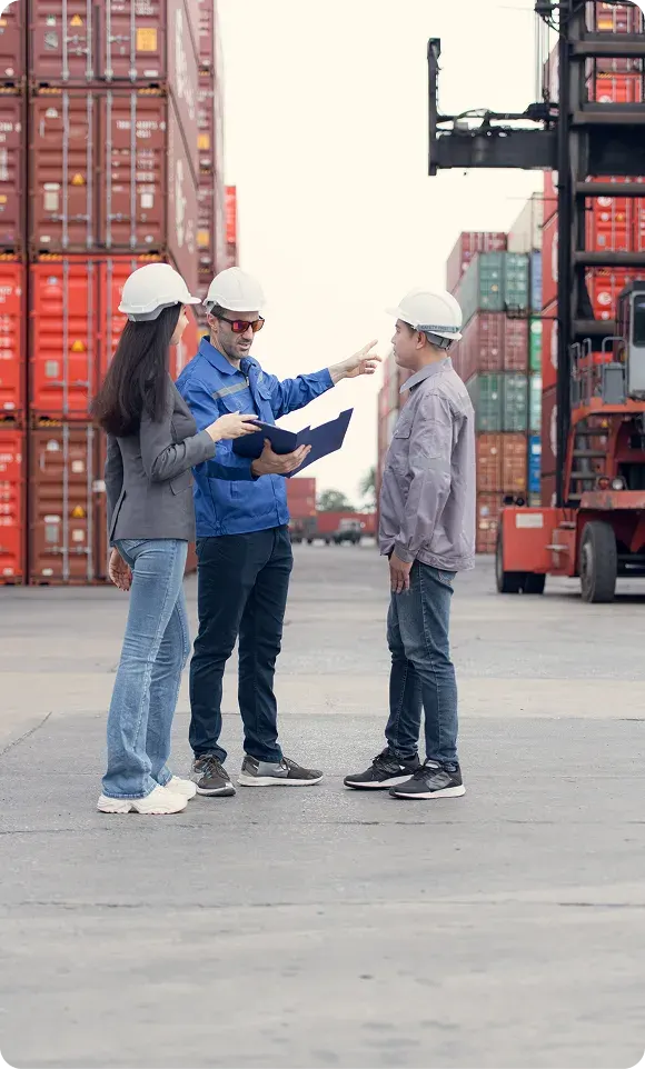 Three workers wearing hard hats discussing plans in front of stacked shipping containers at a port.