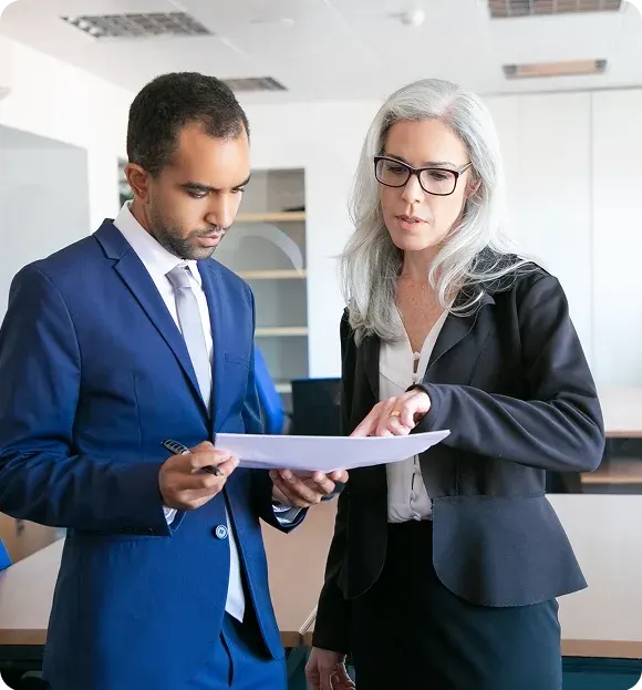 A businesswoman with gray hair and glasses discussing documents with a man in a blue suit in an office.