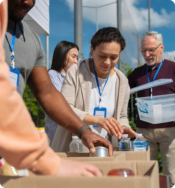 Volunteers wearing name tags sorting and packing canned goods and bottled water outdoors.