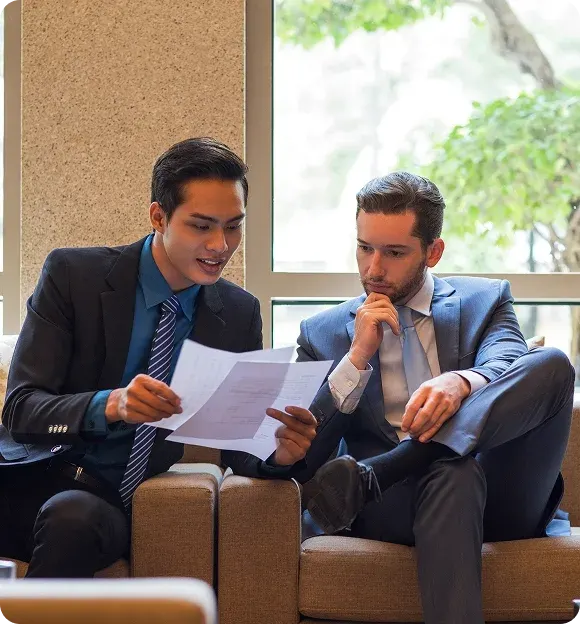 Two businessmen in suits sitting on a couch reviewing documents together near a window.