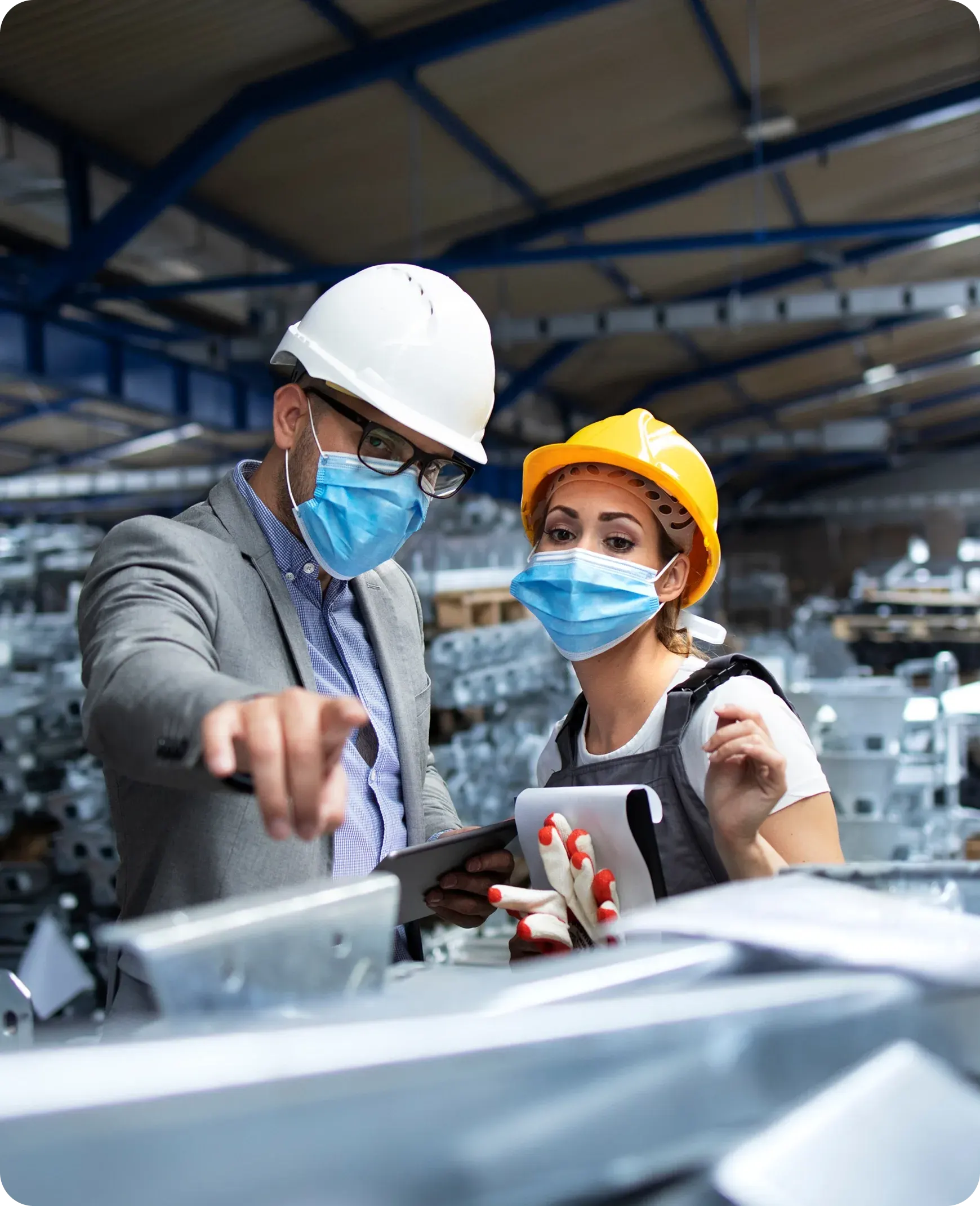 Man and woman wearing safety helmets and masks inspecting metal parts inside a factory.