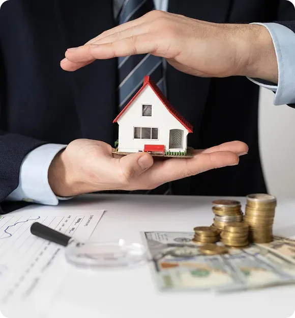 Person in suit holding a small house model between their hands with stacks of coins and dollar bills on a desk.