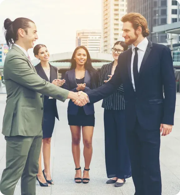 Two businesspeople shaking hands outdoors with three colleagues smiling and clapping behind them in a city setting.