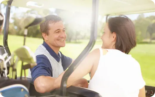 Two people smiling and sitting in a golf cart on a sunny golf course.