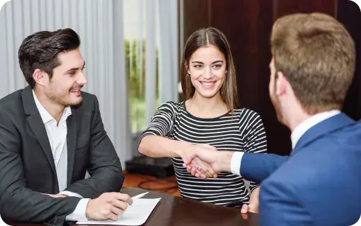 A young woman smiling and shaking hands with a man in a blue suit across a table, while another man in a black suit watches and holds a pen.