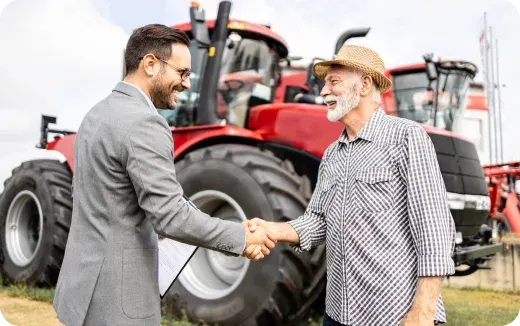 Man in a suit shaking hands with an older man wearing a straw hat and plaid shirt in front of red tractors.