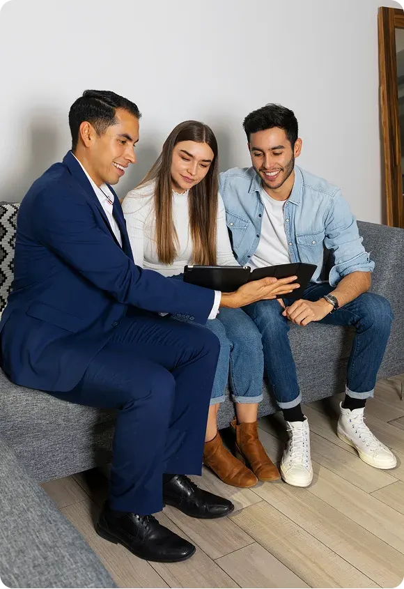 Three people sitting on a gray couch, looking at a tablet with interest and smiling.