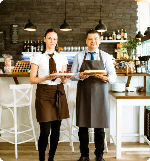 A man and a woman wearing aprons and holding cakes standing in a cozy café with bar stools and a brick wall background.