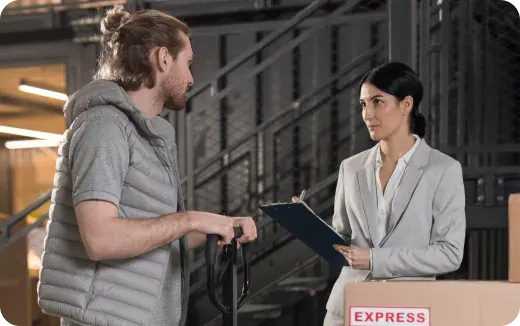 A woman in a light gray blazer holding a clipboard talks to a man in a gray vest with a rolling suitcase in an industrial-style setting with an express shipping box nearby.