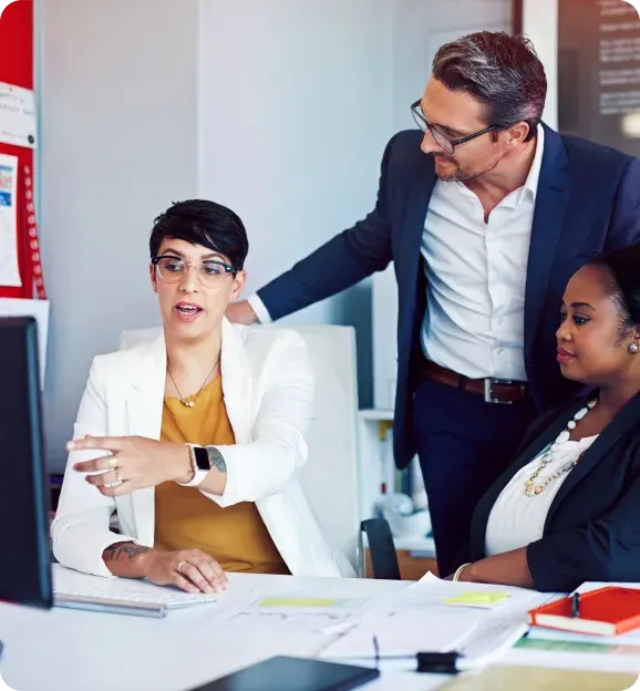 Three business professionals engaged in a discussion at an office desk with documents and a computer screen.
