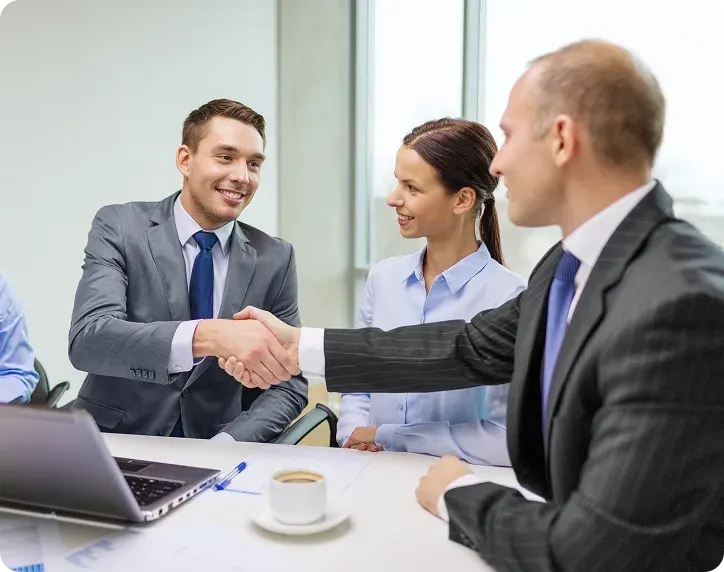 Two businessmen shaking hands across a desk with a businesswoman smiling and watching, in a modern office setting.