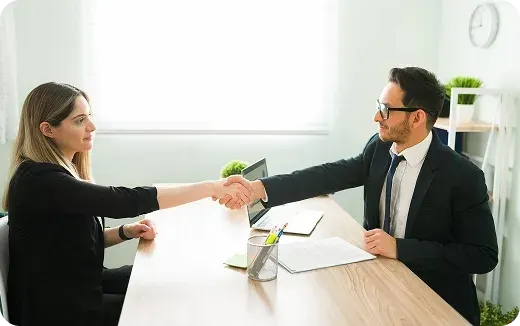 Two business professionals shaking hands across a wooden desk in a bright office.