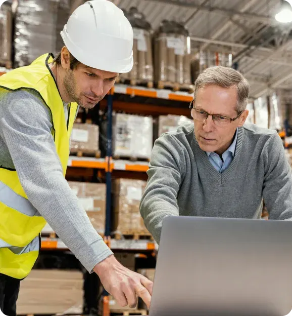 Two men in a warehouse looking at a laptop, one wearing a white hard hat and yellow safety vest, the other in glasses and a gray sweater.