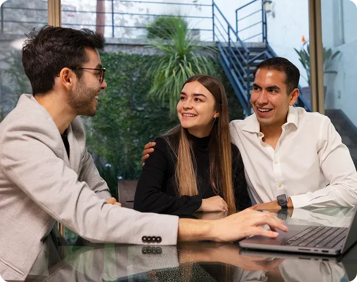 Three young professionals sitting at a glass table, with one using a laptop and the others smiling and engaging in conversation in a modern indoor setting.