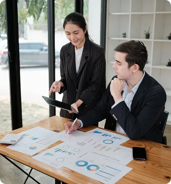 Two business professionals analyzing charts and graphs at a wooden table, one seated and writing while the other stands holding a tablet.