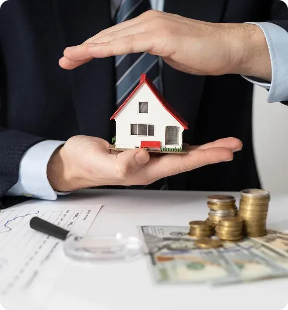 Person in suit holding a small model house with one hand underneath and the other hand protecting it, with coins and dollar bills on a table nearby.