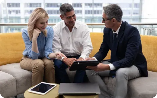 Three professionals sitting on a couch reviewing a tablet with a laptop and another tablet on a small table nearby.