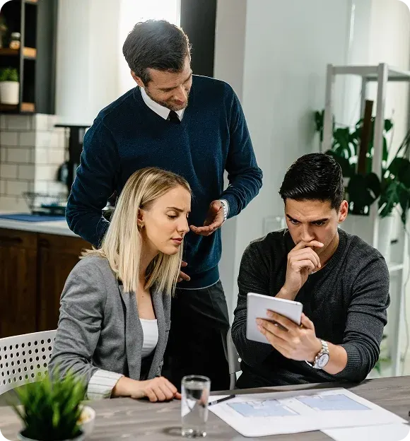 Three coworkers collaborating in an office, with one man standing and explaining while two seated colleagues review information on a tablet.